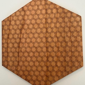 May include: A wooden, hexagonal wall art piece with a honeycomb pattern. The artwork is made of light brown wood and features a repeating design of engraved hexagons. The piece is mounted on a white wall.