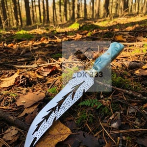 May include: A custom-made knife with a light blue handle and a silver blade. The blade features a unique design with cut-out patterns and dotted accents. The knife is displayed outdoors on a bed of leaves and forest floor.