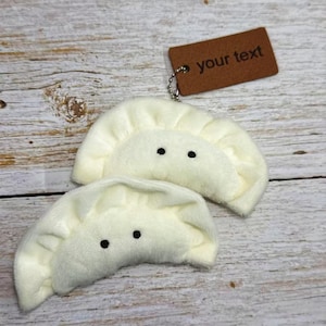 Puede incluir: Dos llaveros de peluche con forma de empanadilla de color crema con ojos de puntos negros. Una etiqueta marrón con el texto "your text" cuelga de una cadena sujeta a una empanadilla. Las empanadillas están sobre una superficie de madera clara.