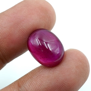 Può includere: Una gemma di rubino lucidata, di forma ovale. La gemma è di un colore rosso-rosa intenso e saturo, con una superficie liscia e riflettente. Il rubino è tenuto tra due dita su uno sfondo bianco, mostrando la sua chiarezza e il suo colore.