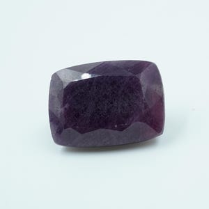 May include: A deep purple, cushion-cut gemstone with multiple facets. The rectangular gem has rounded corners, reflecting light across its surface. The stone is set against a plain white background.