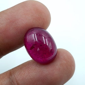 Può includere: Una gemma di rubino lucidata, di forma ovale. La gemma rosso scuro è tenuta tra due dita, mostrando la sua superficie liscia e riflettente. Il colore del rubino è un rosso ricco e saturo, con sottili variazioni di tonalità.