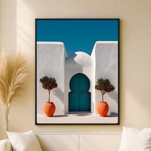 Peut inclure: Impression d'art encadrée représentant un bâtiment blanc avec une porte arquée turquoise et un ciel bleu clair. Deux pots en terre cuite avec de petits arbres encadrent la porte. L'œuvre est présentée dans un cadre noir.