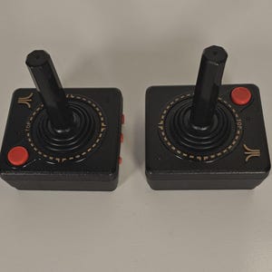 Könnte beinhalten: Zwei schwarze Atari-Joysticks mit roten Knöpfen und einem schwarzen Joystick. Die Joysticks haben ein kreisförmiges Design mit dem Wort "TOP" und dem Atari-Logo. Die Joysticks befinden sich auf einer hellgrauen Oberfläche.