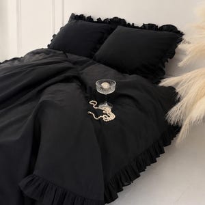 May include: A black bedding set featuring a duvet cover and two matching pillowcases, all adorned with ruffled edges. A clear glass pedestal bowl containing a single silver sphere and a strand of white faux pearls rests on the duvet cover.