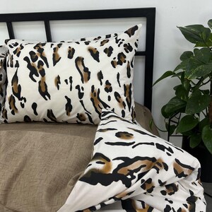 May include: A rectangular pillowcase and a folded duvet cover featuring a white base with a black and brown leopard print pattern. The bedding is displayed on a bed frame with a solid brown textured sheet visible underneath the patterned fabric.
