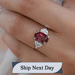 Puede incluir: Un anillo plateado con una gran gema roja oscura de talla ovalada, flanqueada por gemas blancas más pequeñas en un diseño triangular. El anillo se presenta sobre un fondo suave, resaltando el brillo y la elegancia de la joya.