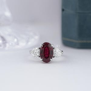 May include: A silver-toned ring featuring a central oval-cut deep red ruby gemstone held by four prongs, flanked by two pear-cut clear white diamonds on either side, set on a polished metal band with a small SD logo mark on the side.
