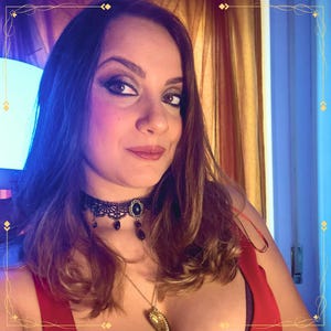 May include: A black lace choker necklace featuring a central oval pendant with a dark stone and small dangling black beads, worn with a gold-toned chain necklace that has a heart-shaped pendant resting against a red garment.