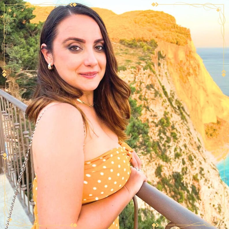 May include: A mustard yellow strapless top featuring a white polka dot pattern, worn with a thin gold chain necklace and small gold hoop earrings, captured in a portrait view with a coastal cliffside landscape in the background.