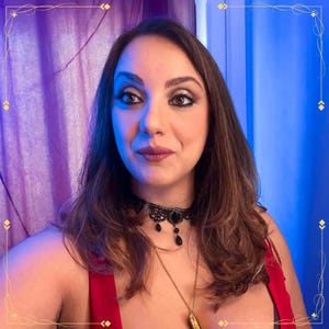 May include: A black lace choker necklace featuring a central oval pendant and small dangling black teardrop beads, worn with a thin gold chain necklace and a red garment against a purple and blue background.