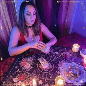 May include: A deck of tarot cards with a floral patterned cloth, a small metal bowl containing stones, and lit candles. The cards feature illustrated figures and symbols, while the dark fabric displays a detailed rose and vine print.