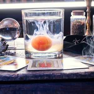 May include: A clear glass tumbler filled with water contains a suspended orange sphere surrounded by swirling white ink. The glass sits on a wooden surface alongside a crystal ball, a glass jar of dried herbs, and several tarot cards.