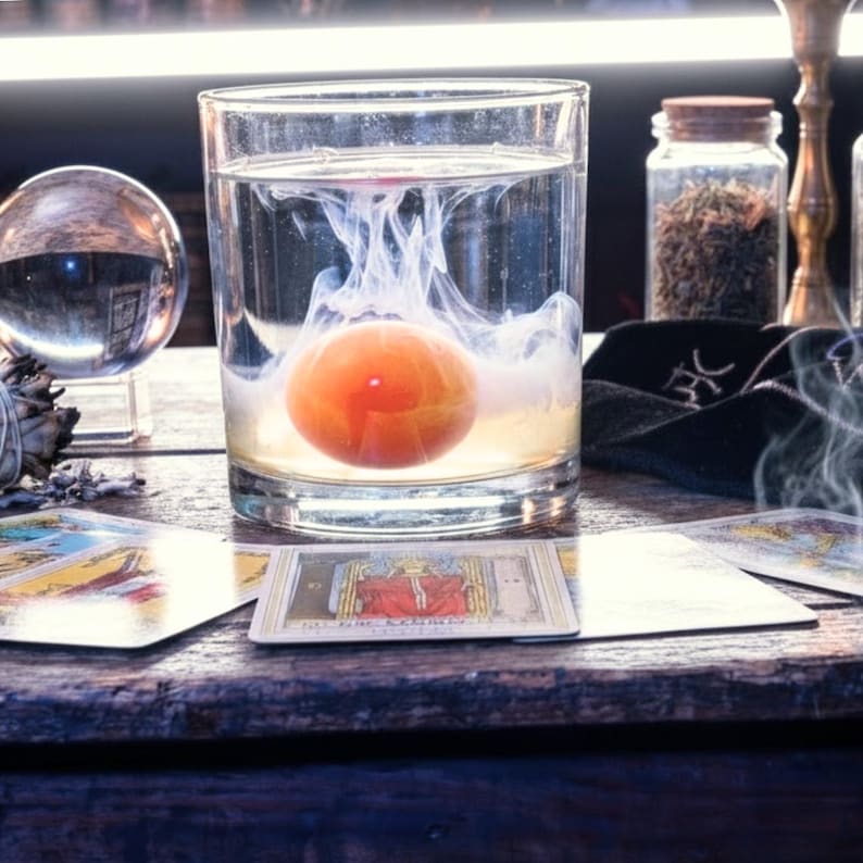 May include: A clear glass tumbler filled with water contains a suspended orange sphere surrounded by swirling white ink. The glass sits on a wooden surface alongside a crystal ball, a glass jar of dried herbs, and several tarot cards.