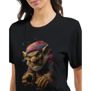 Könnte beinhalten: Schwarzes T-Shirt mit einer Illustration eines Trolls. Der Troll hat ein grimmiges Gesicht, eine große Nase und buntes Fell in Rosa, Blau und Gelb. Das Design ist mittig auf dem T-Shirt platziert.