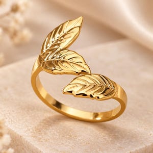 Può includere: Un anello aperto color oro caratterizzato da tre foglie in metallo lucido disposte lungo la fascia. Il design presenta una superficie liscia e riflettente con dettagliate venature incise su ogni singola foglia.