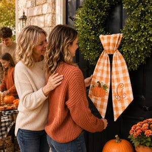 A pumpkin spice orange and white gingham wreath sash features two embroidered designs near the lower ends of the ribbon tails.
One side displays an orange pumpkin with a brown stem, green leaf, and curling vine details.
The opposite side features an elegant monogram letter “G” embroidered in matching orange thread.
Orange, brown, and green threads create a coordinated fall-themed stitched finish.