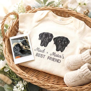 May include: A cream-coloured short-sleeve baby bodysuit featuring an embroidered design of two black dog heads and the text "Mack + Mater's BEST FRIEND" in grey thread, accompanied by a small square photograph of two dogs and two knit booties.