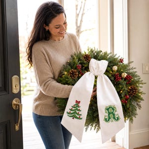 White wreath sash featuring embroidered Christmas designs near the lower ends of both hanging tails.
One side displays a green Christmas tree with red ornaments, a red bow, and a brown trunk, stitched in green, red, and brown threads.
The opposite side features an ornate personalized monogram initial embroidered in layered dark and light green threads.