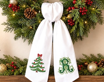 May include: A white hanging towel featuring two embroidered designs: a green Christmas tree with red ornaments and a red bow at the top, and a decorative green monogram letter R with intricate swirling patterns on the right side.