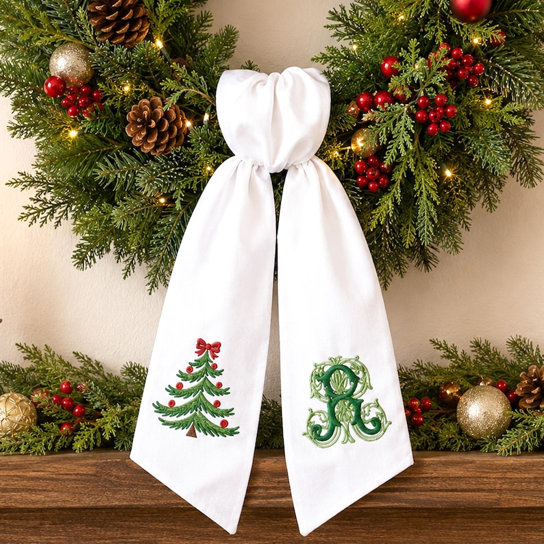 White wreath sash featuring embroidered Christmas designs near the lower ends of both hanging tails.
One side displays a green Christmas tree with red ornaments, a red bow, and a brown trunk, stitched in green, red, and brown threads.
The opposite side features an ornate personalized monogram initial embroidered in layered dark and light green threads.
