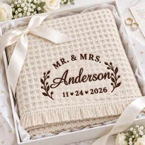 Puede incluir: Una manta de tejido gofrado color crema con flecos, bordada en marrón oscuro con el texto MR. & MRS. Anderson y la fecha 11-24-2026, acentuada con un motivo de hojas decorativo y atada con una cinta de satén crema en una caja de regalo.
