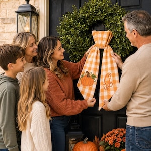 A pumpkin spice orange and white gingham wreath sash features two embroidered designs near the lower ends of the ribbon tails.
One side displays an orange pumpkin with a brown stem, green leaf, and curling vine details.
The opposite side features an elegant monogram letter “G” embroidered in matching orange thread.
Orange, brown, and green threads create a coordinated fall-themed stitched finish.