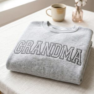 A high-angle view of a folded ash grey sweatshirt placed on a linen-covered table next to a coffee mug and a small vase. The focal point is the large "GRANDMA" embroidery in gray thread, presented in a clean, minimalist aesthetic perfect for a premium product listing.