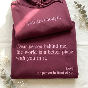 以下が含まれることがあります： 2つのマルーン色のパーカーが重ねられており、それぞれに白い文字が刺繍されています。上のパーカーには「you are enough.」と書かれています。下のパーカーには「Dear person behind me, the world is a better place with you in it. Love, the person in front of you.」と書かれています。