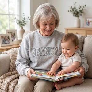 A heartwarming shot of a grandmother sitting on a couch, wearing the ash grey "GRANDMA" embroidered sweatshirt, reading a book to a baby on her lap. The soft studio lighting captures the clean texture of the fabric and the precision of the grey varsity-style embroidery.