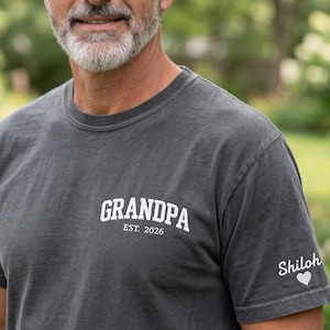 May include: A charcoal gray crew neck t-shirt featuring white screen-printed text on the chest that reads GRANDPA EST. 2026. The left sleeve displays the name Shiloh in white script lettering accompanied by a small white heart icon.