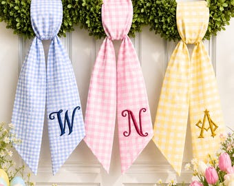 May include: Three fabric ribbons in light blue, pink, and yellow gingham patterns, each featuring a unique embroidered monogram letter. The blue ribbon displays a dark blue W, the pink ribbon a dark pink N, and the yellow ribbon a gold A.