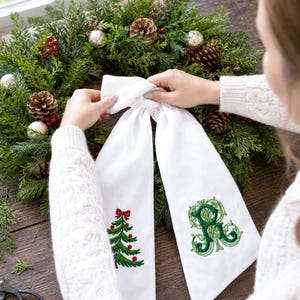 White wreath sash featuring embroidered Christmas designs near the lower ends of both hanging tails.
One side displays a green Christmas tree with red ornaments, a red bow, and a brown trunk, stitched in green, red, and brown threads.
The opposite side features an ornate personalized monogram initial embroidered in layered dark and light green threads.