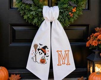May include: A white fabric door hanger featuring a ghost wearing a witch hat, holding a broom and a jack-o'-lantern, with a large orange embroidered letter M on the other side. The hanger is draped over a green wreath with orange flowers.
