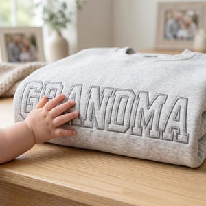 A folded ash grey sweatshirt on a wooden surface with a baby’s hand gently touching the embroidered "GRANDMA" text. The collegiate-style lettering is embroidered in a matching gray thread, highlighting the high-quality craftsmanship and the sentimental, family-oriented theme.