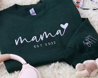 Könnte beinhalten: Ein dunkelgrünes Sweatshirt mit dem Wort "Mama" in weißer Schrift, einem weißen Herz und "EST 2022". Der Ärmel zeigt einen Namen und ein Bogendesign. Das Sweatshirt liegt auf einer weißen, flauschigen Oberfläche.