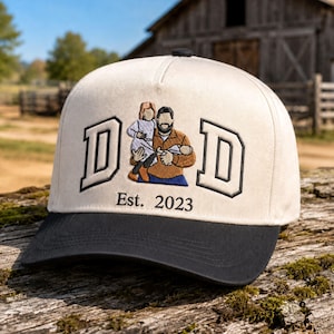 May include: A cream-colored baseball cap with a black brim featuring an embroidered design of a person holding a child and an infant. The word DAD is stitched in large black-outlined letters with the text Est. 2023 centered below the figures.