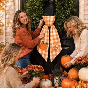 A pumpkin spice orange and white gingham wreath sash features two embroidered designs near the lower ends of the ribbon tails.
One side displays an orange pumpkin with a brown stem, green leaf, and curling vine details.
The opposite side features an elegant monogram letter “G” embroidered in matching orange thread.
Orange, brown, and green threads create a coordinated fall-themed stitched finish.
