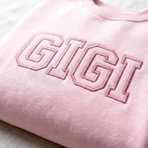 A close-up shot of a light pink sweatshirt featuring a large, centered embroidery of the word "GIGI" in a classic varsity block font. The design is meticulously stitched with deep rose thread, creating a sophisticated tone-on-tone effect with a textured, premium finish.