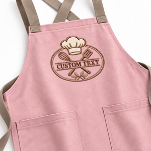 May include: A light pink apron with brown cross-back straps and two front pockets. The chest features an embroidered brown oval emblem containing a chef hat, a whisk, a spatula, and the text CUSTOM TEXT centered in the middle.