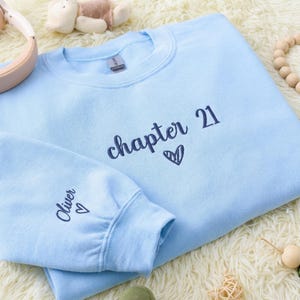 Puede incluir: Sudadera azul claro con las palabras "chapter 21" y un corazón. La manga tiene el nombre "Oliver" y un corazón. La sudadera está hecha de un material suave.