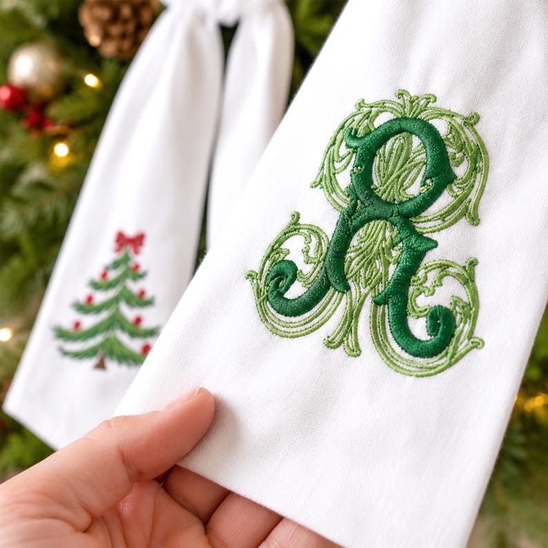 White wreath sash featuring embroidered Christmas designs near the lower ends of both hanging tails.
One side displays a green Christmas tree with red ornaments, a red bow, and a brown trunk, stitched in green, red, and brown threads.
The opposite side features an ornate personalized monogram initial embroidered in layered dark and light green threads.
