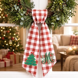 May include: A red and white buffalo check fabric ribbon hanging from a green wreath. The left tail features an embroidered green Christmas tree with red ornaments, while the right tail displays an embroidered green letter M with leafy accents.