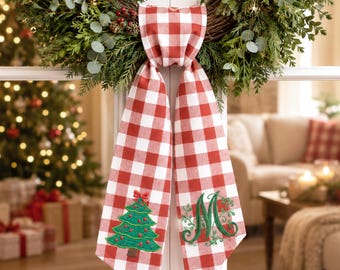 May include: A red and white buffalo check fabric ribbon hanging from a green wreath. The left tail features an embroidered green Christmas tree with red ornaments, while the right tail displays an embroidered green letter M with leafy accents.