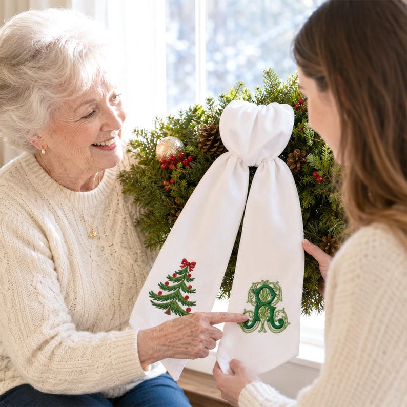 White wreath sash featuring embroidered Christmas designs near the lower ends of both hanging tails.
One side displays a green Christmas tree with red ornaments, a red bow, and a brown trunk, stitched in green, red, and brown threads.
The opposite side features an ornate personalized monogram initial embroidered in layered dark and light green threads.