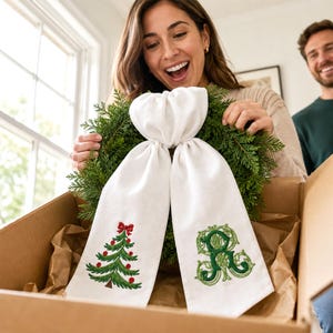 White wreath sash featuring embroidered Christmas designs near the lower ends of both hanging tails.
One side displays a green Christmas tree with red ornaments, a red bow, and a brown trunk, stitched in green, red, and brown threads.
The opposite side features an ornate personalized monogram initial embroidered in layered dark and light green threads.