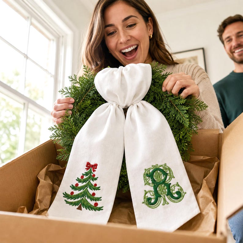 White wreath sash featuring embroidered Christmas designs near the lower ends of both hanging tails.
One side displays a green Christmas tree with red ornaments, a red bow, and a brown trunk, stitched in green, red, and brown threads.
The opposite side features an ornate personalized monogram initial embroidered in layered dark and light green threads.