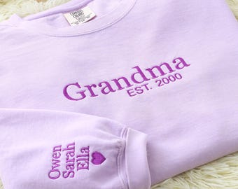 May include: A light purple sweatshirt with the word "Grandma" embroidered in purple thread, along with "EST. 2000". The sleeve features embroidered names and a heart. The sweatshirt is made of a soft material.