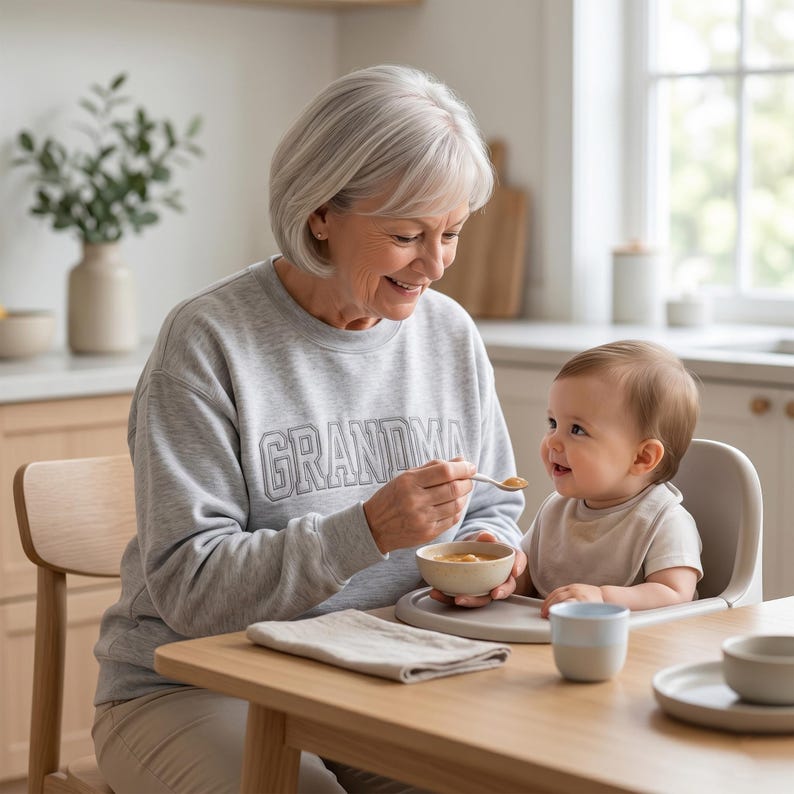 A lifestyle image of an elderly woman wearing an ash grey sweatshirt with "GRANDMA" embroidered in gray thread while feeding a baby in a high chair. The scene is set in a bright, modern kitchen, emphasizing the comfort and daily wearability of the personalized apparel.