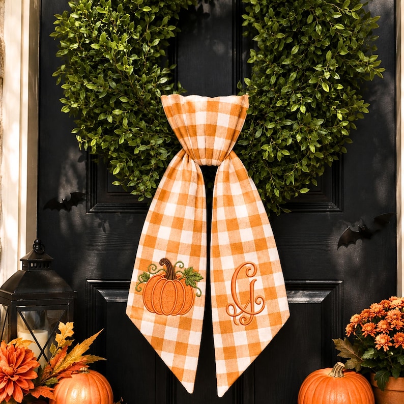 A pumpkin spice orange and white gingham wreath sash features two embroidered designs near the lower ends of the ribbon tails.
One side displays an orange pumpkin with a brown stem, green leaf, and curling vine details.
The opposite side features an elegant monogram letter “G” embroidered in matching orange thread.
Orange, brown, and green threads create a coordinated fall-themed stitched finish.